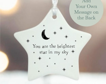 May include: White ceramic star-shaped ornament with a crescent moon and stars printed on it. The ornament reads "You are the brightest star in my sky". The back of the ornament is blank and can be personalized with a message.