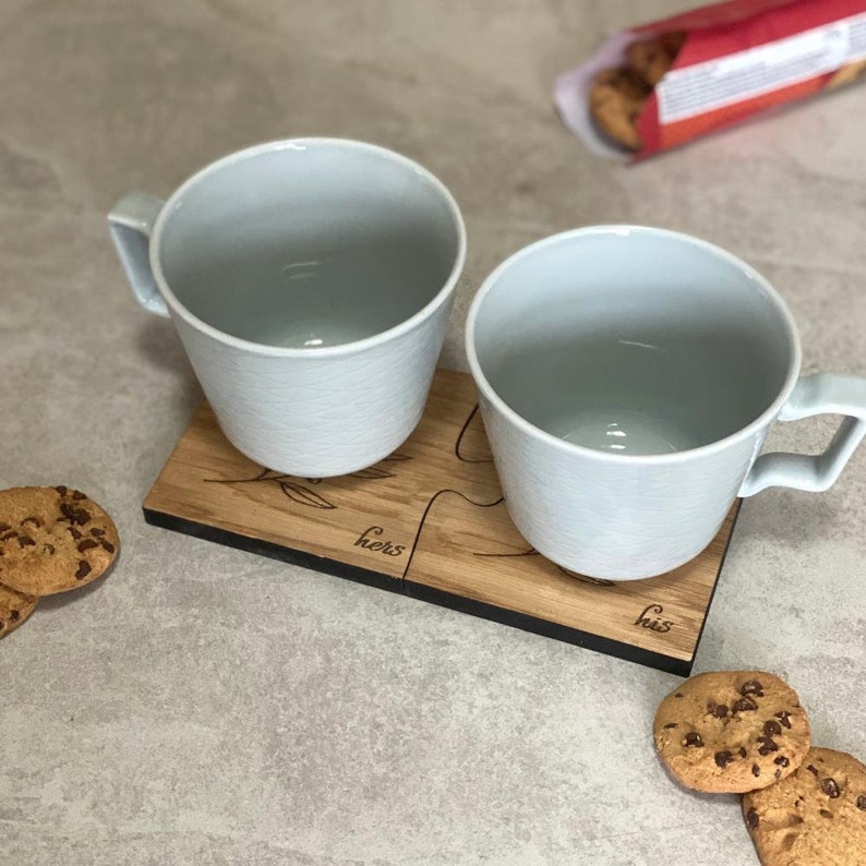May include: Two white ceramic mugs on a wooden coaster with the words "his" and "hers" engraved on it. The coaster is made of two pieces that fit together like a puzzle. There are chocolate chip cookies on the table.