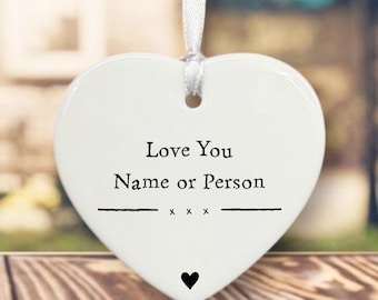 May include: White heart-shaped ornament with the text "Love You Name or Person" in black. A black heart is at the bottom. The ornament hangs from a white ribbon. The background is blurred.