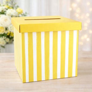 May include: A square box with a removable lid featuring a horizontal slot on top. The exterior of the box is decorated with a vertical striped pattern in bright yellow and white. The box is constructed from a smooth material with a glossy finish.