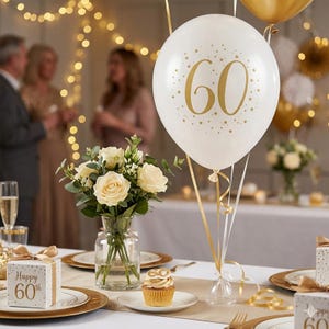 May include: A celebratory table setting for a 60th birthday. A white balloon with gold '60' and star details is central. Other elements include a bouquet of white roses, a cupcake, and gift boxes, all decorated with gold accents.