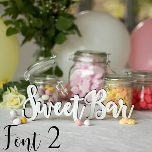 May include: White wooden sign with the text "Sweet Bar" in a cursive font. The sign is sitting on a table with jars of candy in the background. The text "Font 2" is written in black below the sign.