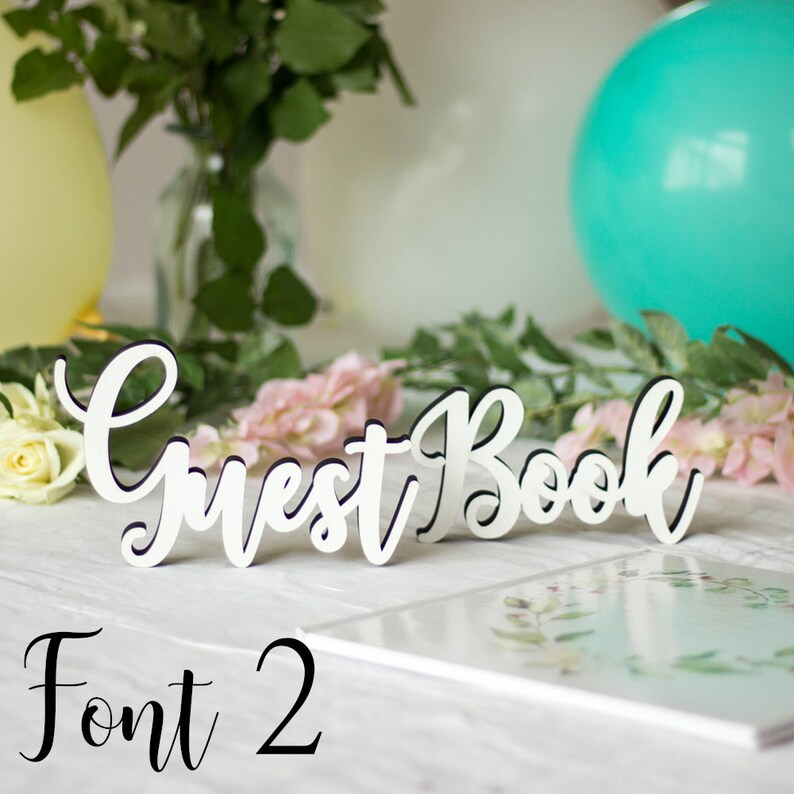 May include: White wooden sign with the words "Guest Book" in a cursive font. The sign is sitting on a white table with a white book and flowers in the background. The text "Font 2" is written below the sign.