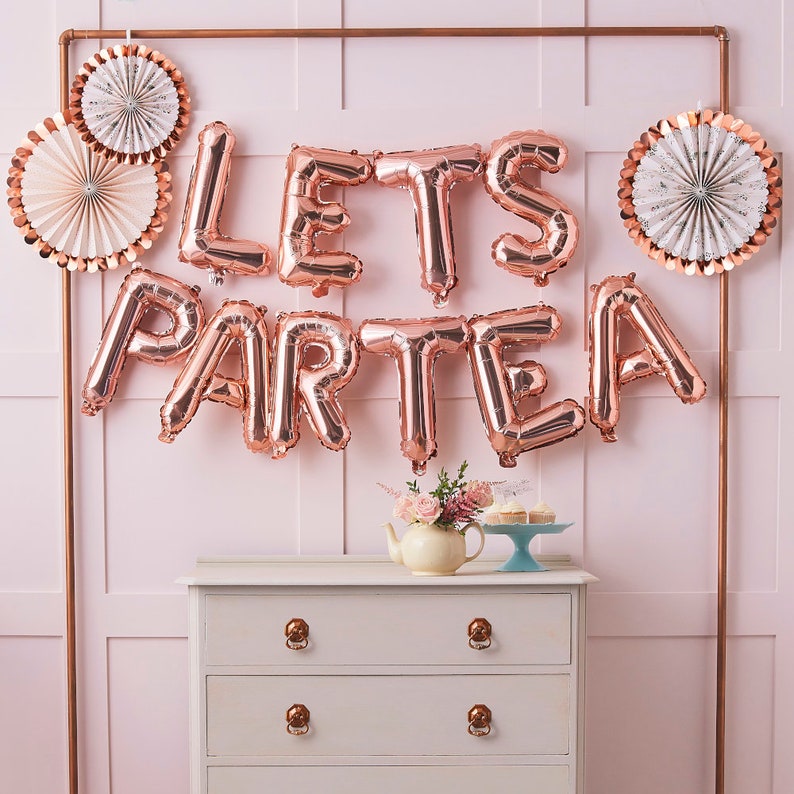 K&ouml;nnte beinhalten: Ros&eacute;goldene Folienballons, die "Let's Partea" buchstabieren, h&auml;ngen an einer rosa Wand mit einer wei&szlig;en Kommode und ros&eacute;goldenen Papierf&auml;chern.
