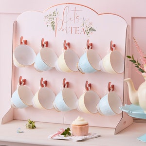 K&ouml;nnte beinhalten: Ein rosa Holzst&auml;nder f&uuml;r Teetassen mit dem Text "Let's ParTEA" in goldenen Buchstaben. Der St&auml;nder hat 10 Haken zum Aufh&auml;ngen von Teetassen, und es h&auml;ngen 10 Teetassen an den Haken. Die Teetassen sind in drei Farben: hellblau, creme und gelb. Auf dem Tisch vor dem St&auml;nder steht ein Cupcake.