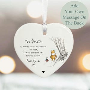 May include: White heart-shaped ceramic ornament with a ribbon for hanging. Features a Winnie the Pooh illustration and text: "Mrs Rossiter, It makes such a difference, said Pooh, To have someone who believes in you, love Cara xx".