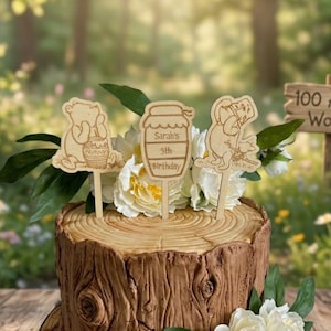 May include: Three wooden cake toppers featuring laser-cut designs of a bear with a honey pot, a honey jar with the text Sarah's 5th Birthday, and a bear with a small animal, all mounted on sticks and placed in a cake decorated like a tree stump.