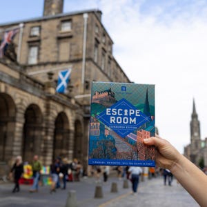 May include: A colourful box for the Escape Room Edinburgh Edition game, featuring illustrations of Edinburgh landmarks. The box displays the text "ESCAPE ROOM" in blue, with additional text below. The game promises 11 puzzles and 60 minutes to escape.