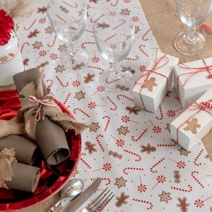 Könnte beinhalten: Eine festliche Weihnachts-Tischdekoration mit einer weißen Tischdecke mit roten Zuckerstangen, Lebkuchenmännern und Sternen. Enthält rote Teller, braune Servietten in Jute gewickelt, klare Weingläser und kleine weiße Geschenkboxen mit roter Schnur und Lebkuchenmann-Akzenten.