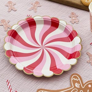 May include: A decorative paper plate featuring a red, pink, and white spiral pattern. The plate has a scalloped edge with a gold trim. Gingerbread man cutouts and a candy cane are scattered around, suggesting a festive theme.
