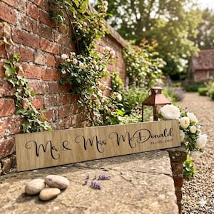 May include: A wooden sign engraved with "Mr & Mrs McDonald" and the date "21.05.2019". The sign is set against a brick wall with climbing plants and a stone ledge. A lantern and flowers are also visible.