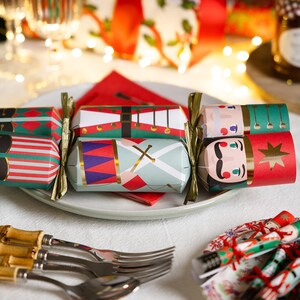 May include: Three cylindrical Christmas crackers featuring nutcracker-themed designs in red, green, white, and gold. The crackers are tied with gold ribbon at the ends and are arranged on a white plate with a red napkin, alongside silver cutlery.