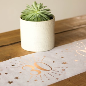 May include: A white table runner with a gold foil design featuring the number 30 surrounded by stars. The runner is on a wooden table with a small potted succulent plant.