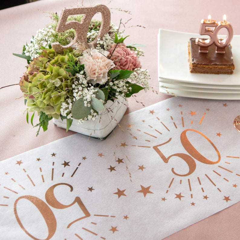 May include: A white table runner with a rose gold design featuring the number 50 with stars and rays of light. A centerpiece of flowers in a white vase with a rose gold 50 on top is on the table. A chocolate cake with a rose gold 50 candle is on a white plate.