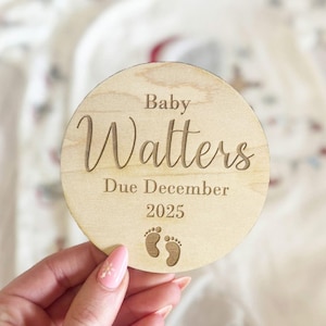 May include: A round wooden sign with the text "Baby Watters Due December 2025" and a pair of baby footprints.