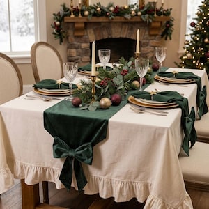 May include: A cream-colored tablecloth with a ruffled edge, featuring a dark green velvet table runner adorned with a large bow at the end. The table is set with gold-rimmed plates, dark green napkins with star napkin rings, and crystal glasses.