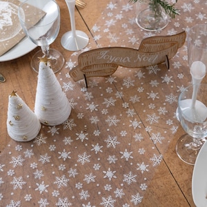 Op de afbeelding: Een feestelijke tafelsetting met witte en gouden accenten. Twee witte kegelvormige kerstbomen met gouden sterren staan naast een houten bord met de tekst "Joyeux Noël". De tafel is versierd met een sneeuwvlokkenpatroon en glaswerk.