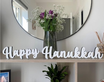 May include: White wooden letters spelling "Happy Hanukkah" on a shelf with a vase of purple flowers and a potted plant.