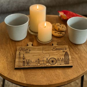 Può includere: Un set di sottobicchieri in legno a forma di puzzle con un design dello skyline di Londra. Il sottobicchiere è inciso con "Matt's Coffee" e "Emma's Tea". Due candele accese, due tazze e biscotti su un tavolo di legno.