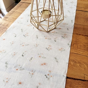 May include: A floral patterned table runner with a light beige background and small orange, white, and green flower illustrations. A gold geometric candle holder with a lit candle sits on top of the runner.