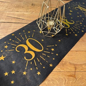 以下が含まれることがあります： 黒のテーブルランナーに金色の星と数字の30が印刷されています。数字の30は金色の光線で囲まれています。ランナーの上に金色の幾何学的なキャンドルホルダーが置かれています。