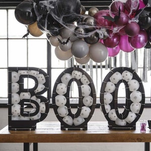 以下が含まれることがあります： 白いクモの巣で飾られた、黒と白の風船文字で「BOO」と綴られています。文字は厚紙製で、白い風船で満たされています。風船は文字の周りに円形に配置されています。文字は木のテーブルの上に置かれています。