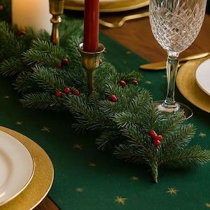 May include: A festive table setting featuring a green table runner adorned with gold stars. A garland of evergreen branches and red berries runs down the center, with a red candle in a gold candlestick. A crystal wine glass and gold-rimmed plates complete the holiday scene.