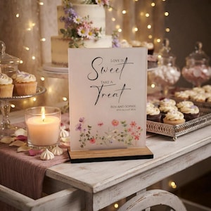 May include: A rectangular acrylic sign with a wooden base featuring the black text "Love is Sweet Take a Treat" and "Ben and Sophie 10th August 2026" above a colourful watercolour illustration of various wildflowers in shades of pink, purple, and green.