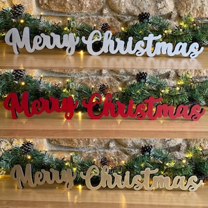 Peut inclure: Trois pancartes en bois "Joyeux Noël" de différentes couleurs : argent, rouge et or. Les pancartes sont disposées sur une étagère en bois avec une guirlande verte et des lumières.
