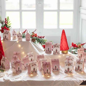 Könnte beinhalten: Eine Sammlung kleiner, hausförmiger Geschenkboxen aus Papier mit rot-weißen vertikalen Streifen und weihnachtlichen Illustrationen. Jede Box hat ein Giebeldach mit einer Kordel und ist auf einer weißen Stoffoberfläche arrangiert.