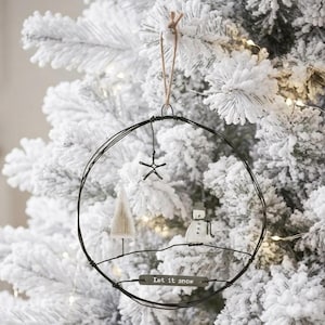 Op de afbeelding: Een cirkelvormig ornament van zwart draad met een kleine witte houten boom, een witte sneeuwpopfiguur, een metalen ster en een rechthoekig metalen label met de tekst Let it snow, opgehangen aan een bruine touwlus.