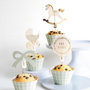 May include: Cupcakes with decorative toppers, including a rocking horse, baby carriage, and bow. The cupcakes are in light green and white checkered cups. One topper reads "OH BABY" in gold lettering. Ideal for a baby shower.
