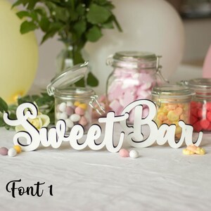 May include: White wooden sign with the text "Sweet Bar" in a cursive font. The sign is sitting on a white table with jars of candy in the background.