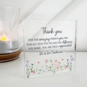 Może przedstawiać: Przezroczysty akrylowy blok z napisem "Thank you" elegancką czcionką. Tekst kontynuuje się z przesłaniem wdzięczności. Na dole znajduje się kwiatowy wzór z różowymi i fioletowymi kwiatami.