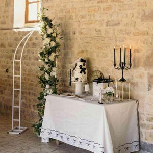 May include: An elegant wedding reception setup featuring a white tablecloth with black bow detailing. A tiered cake, decorated with black flowers and a bow, is displayed on a stand. Black candlesticks and a floral arch add to the refined atmosphere.
