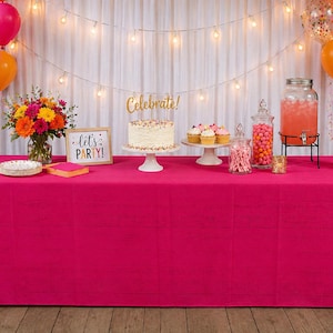 May include: A party table with a bright pink tablecloth featuring a white cake with a gold Celebrate topper, cupcakes, a glass beverage dispenser with pink liquid, jars of sweets, a bouquet of colourful flowers, and a sign reading Let's PARTY!
