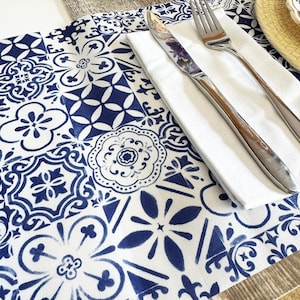 Könnte beinhalten: Eine weiß-blaue Tischdecke mit floralem und geometrischem Muster. Eine gefaltete weiße Serviette, eine Gabel und ein Messer liegen auf der Tischdecke. Das Besteck ist aus Edelstahl. Die Tischdecke liegt auf einer Holzoberfläche.