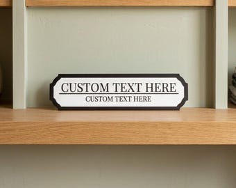 May include: A rectangular sign with the text "CUSTOM TEXT HERE" in a black and white design. The sign is on a wooden shelf, with a light green wall in the background. Other items are on the shelf, including a clock and books.