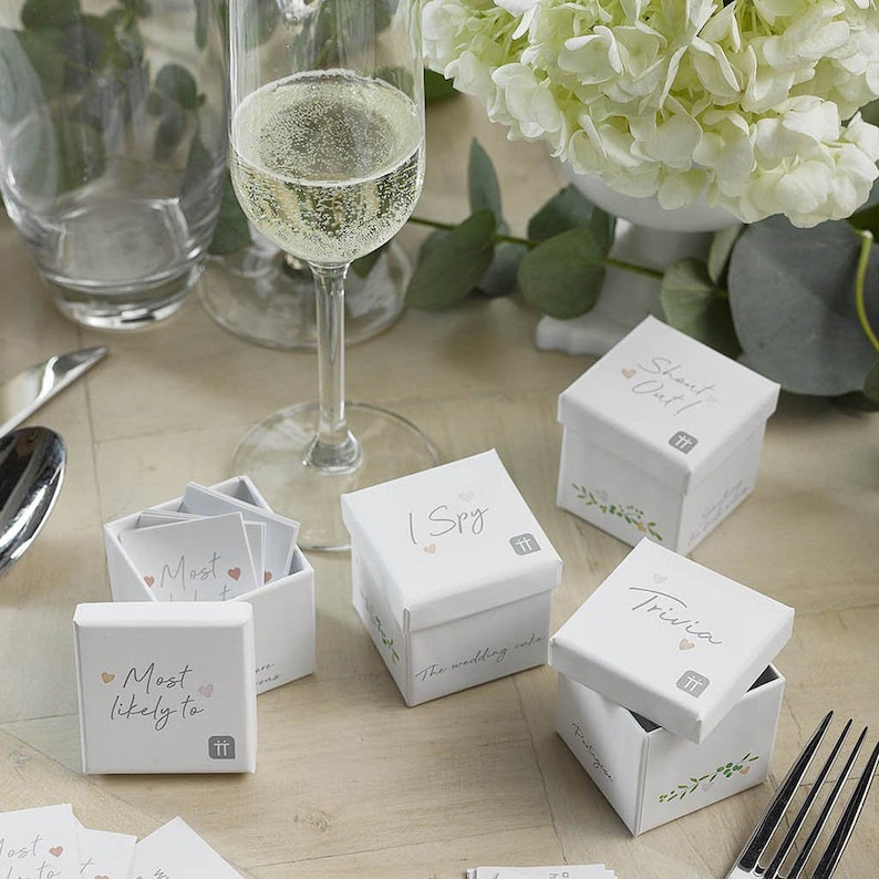 May include: White boxes with floral designs and the words "I Spy", "Most Likely To", "Shout Out!" and "Trivia" on them. The boxes are filled with small cards.