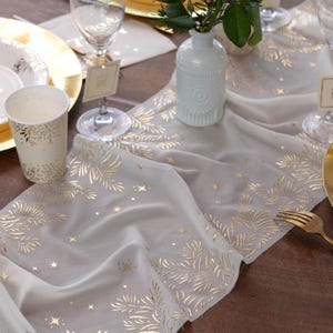 May include: A pale grey table runner featuring a gold leaf and star design. The runner is laid on a brown wooden table, with a white vase, gold-edged plates, and a paper cup with a gold leaf pattern. A gold fork and wine glasses are also present.