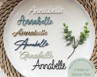 Peut inclure: Six découpes personnalisées du prénom Annabelle dans différentes couleurs et polices, disposées sur une assiette blanche à côté d'un petit brin vert. Un autocollant circulaire sur l'assiette indique : Over 45 colours available, 6 fonts to choose from.