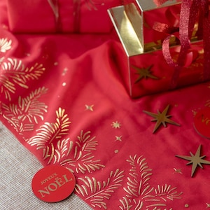 Op de afbeelding: Een feestelijke rode stof met gouden dennenboom- en sterpatronen. Een rode cirkel met de tekst "JOYEUX NOËL" is zichtbaar. Gouden geschenkdozen en stervormige ornamenten dragen bij aan het feestelijke thema.