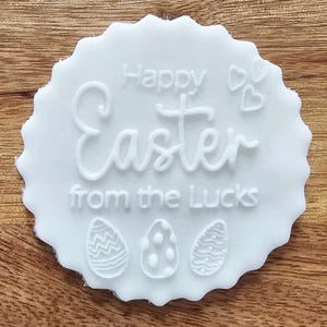 May include: A white, scalloped-edge Easter decoration with the words "Happy Easter from the Lucks" in a cursive font. The design includes three Easter eggs and two small hearts.