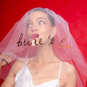 Op de afbeelding: Een doorschijnende witte bruidssluier met gouden folieletters met de tekst "bride to be". De sluier is over het hoofd van een persoon gedrapeerd, tegen een rode achtergrond. De persoon draagt een witte jurk en een gouden ketting.