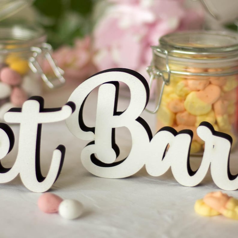 Puede incluir: Letras de madera blancas que deletrean "Sweet Bar" con contornos negros. Las letras est&aacute;n sobre una mesa blanca con tarros de dulces en el fondo.