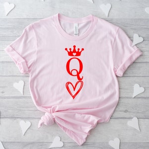 May include: Pink t-shirt with a red graphic of a queen of hearts playing card with a crown and a heart.
