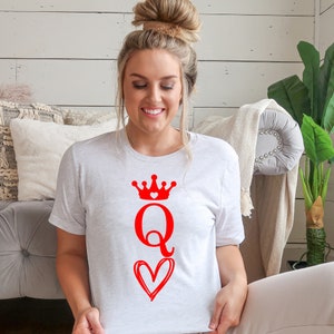May include: A white t-shirt with a red queen of hearts design. The design features a crown with a small heart in the center, the letter Q, and a heart with a swirl design.