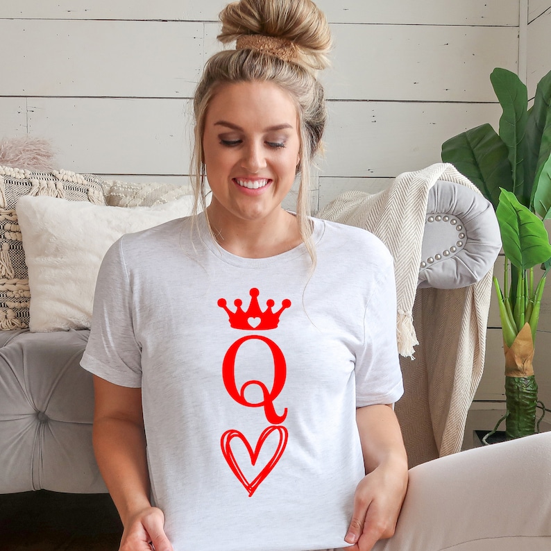 May include: A white t-shirt with a red queen of hearts design. The design features a crown with a small heart in the center, the letter Q, and a heart with a swirl design.