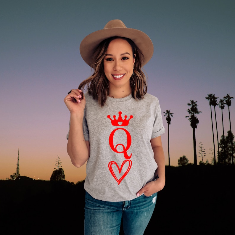 May include: A woman wearing a gray t-shirt with a red queen of hearts graphic. The graphic features a red queen with a crown and a heart.