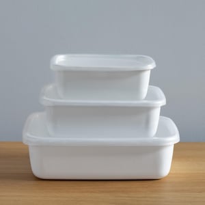 以下が含まれることがあります： 透明な蓋が付いた、白の長方形の食品保存容器が3つ重ねられています。容器は、一番大きいものが一番下になるように積み重ねられ、木製の表面に置かれています。背景には水色の壁があります。